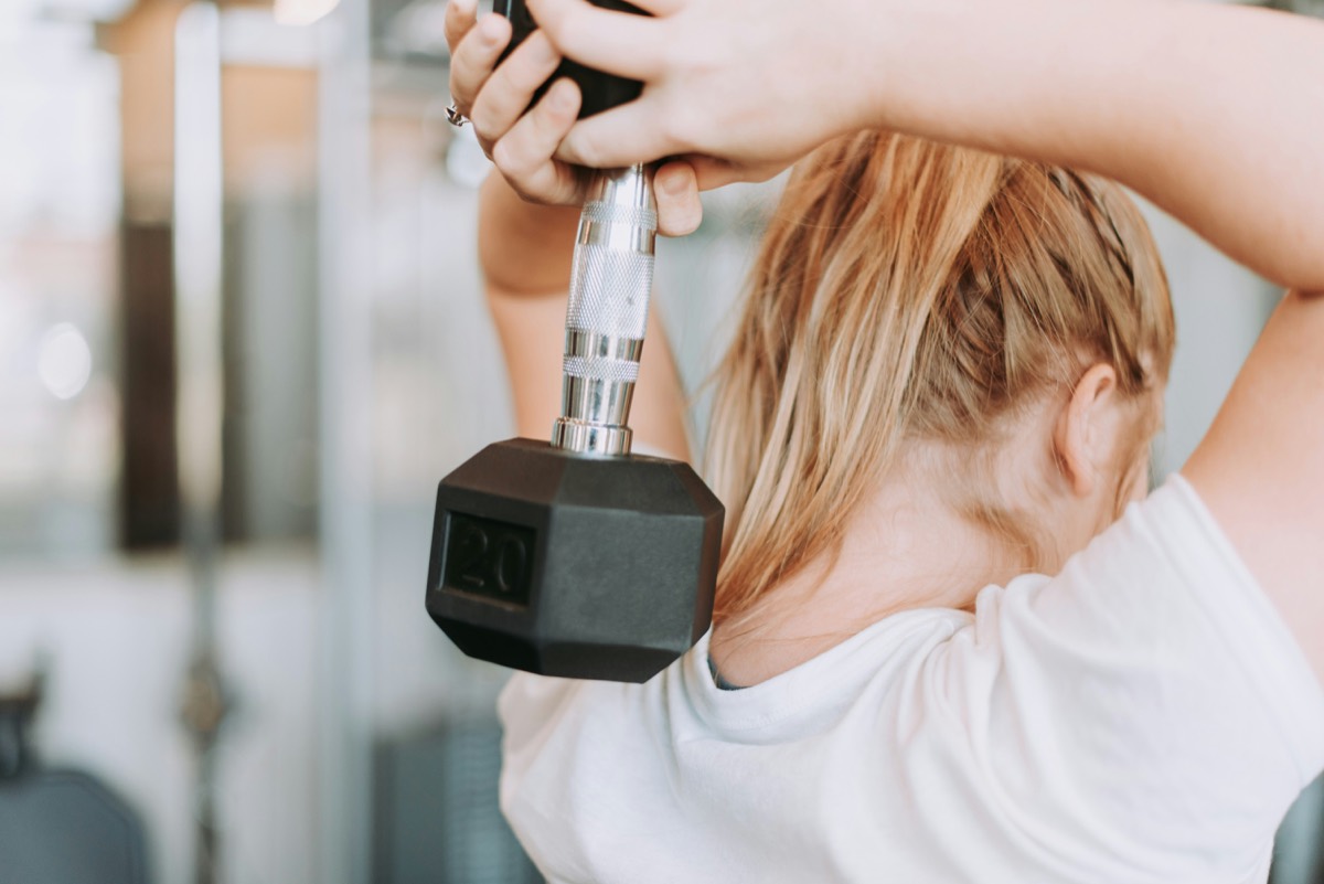 A woman performing a dumbbell triceps extension in a gym.